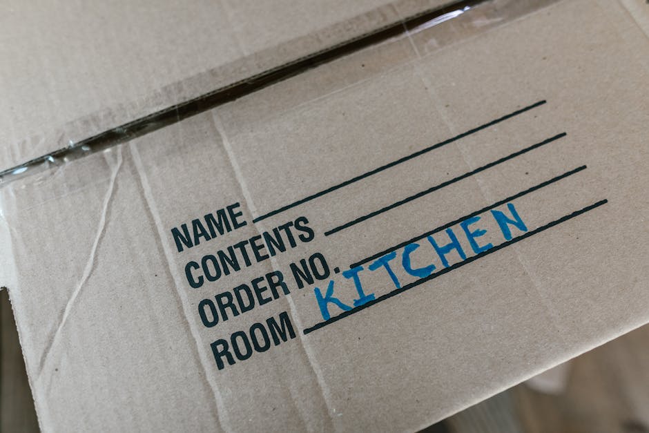 A woman with curly dark hair, dressed in a white t-shirt, blue jeans, and white sneakers, is seated among numerous cardboard moving boxes of various sizes wrapped with red and black tape, inside a roo