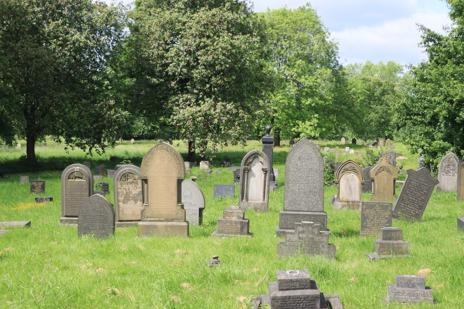 A stone statue group located in a cemetery, depicting three women in classical draped attire, with one woman standing and raising her right arm, another seated holding a small child, and a third woman