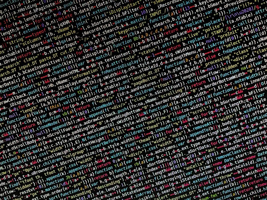 A close-up of a person's hand typing on a laptop keyboard positioned on a wooden surface, with the screen displaying lines of code or text. The person is wearing a wristwatch, and the environment appe